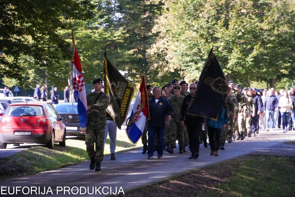Mimohod sudionika, uključujući vojnike u maskirnim uniformama i građane s hrvatskim zastavama i zastavama postrojbi, na sunčanom danu u parku.
