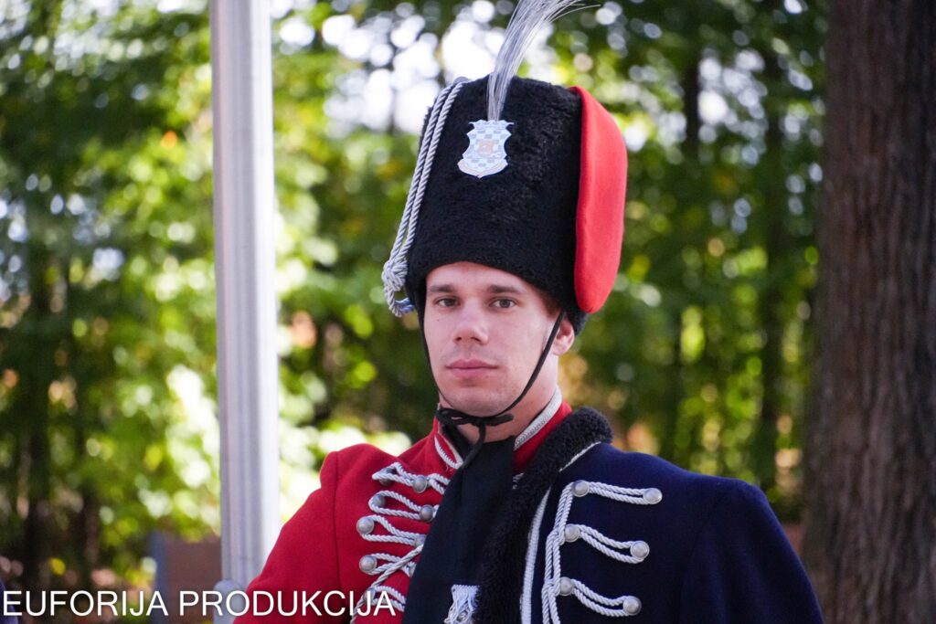 Portret pripadnika povijesne počasne garde u tradicionalnoj crveno-plavoj uniformi s visokom krznenom kapom i perjanicom, na pozadini zelenog drveća.