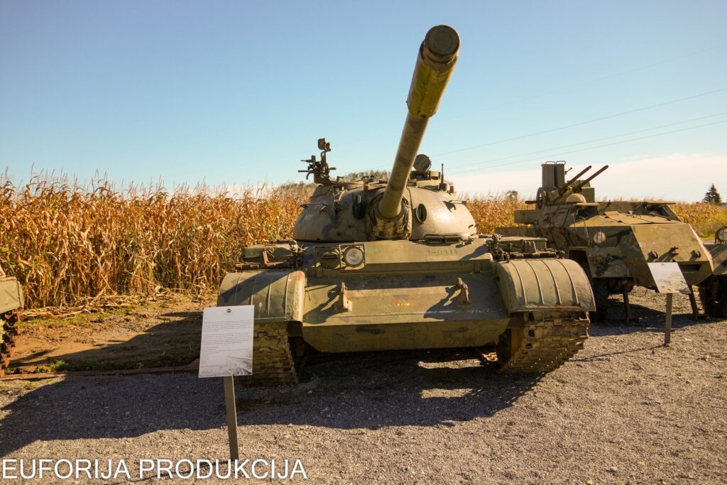 Izložen tenk T-55 i vojna oklopna vozila na Spomen-području Barutana, s poljem kukuruza u pozadini pod sunčanim nebom.